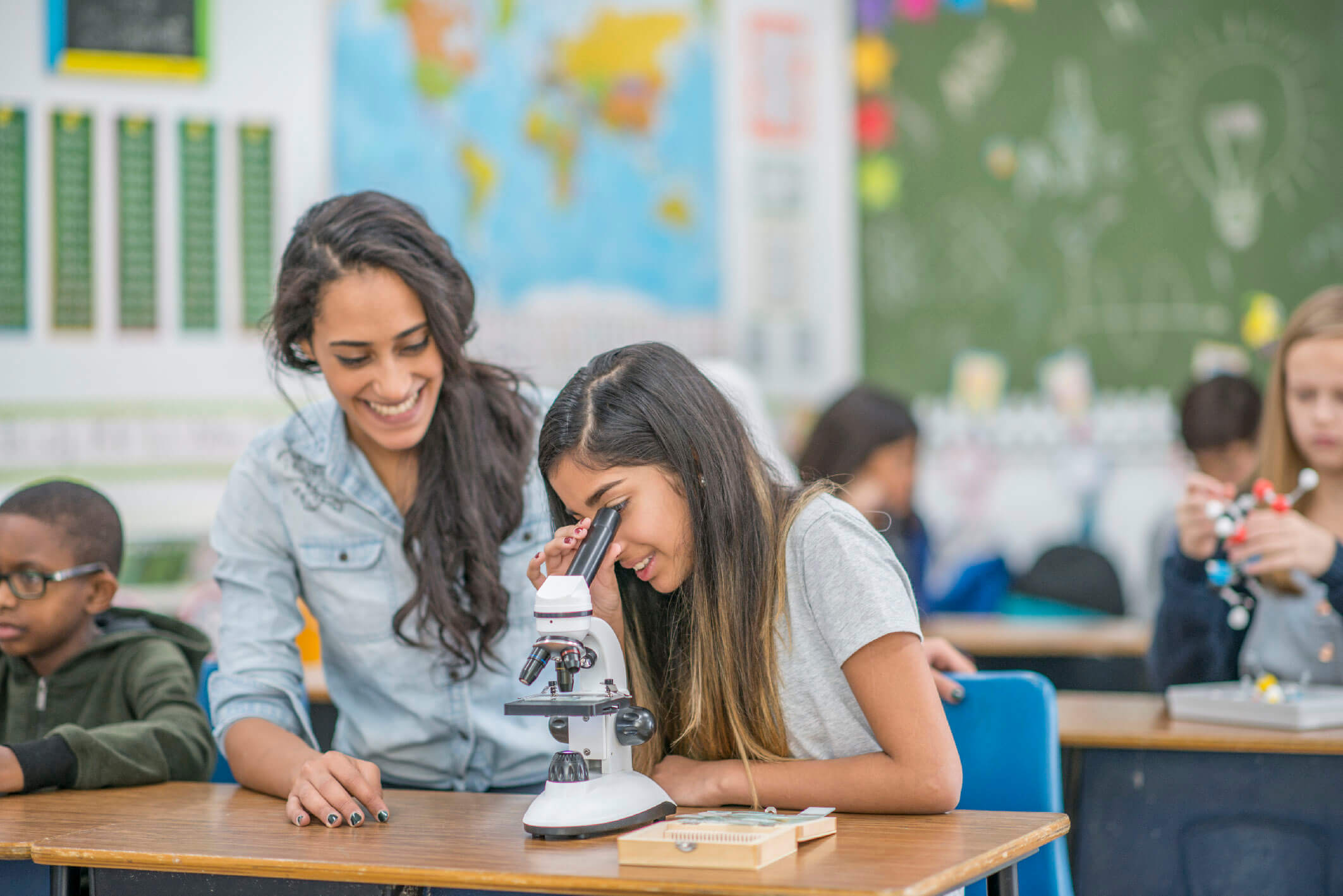 An elementary school girl and her teacher are working together on a science project. The girl is looking through a microscope.